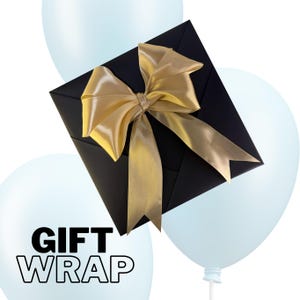 May include: A black gift box with a gold satin bow, set against a backdrop of light blue balloons. The words "GIFT WRAP" are printed in bold, white letters on a balloon. The bow adds a touch of elegance to the presentation.