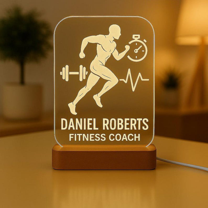 May include: A personalized fitness-themed night light with a wooden base. The light features a running figure, a barbell, a stopwatch, and a heartbeat line. The text "DANIEL ROBERTS FITNESS COACH" is displayed below the graphics.