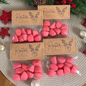 Puede incluir: Una composición navideña festiva con caramelos rosas en bolsas transparentes, etiquetados como "Rentier Nasen", sobre un salvamanteles tejido. Incluye adornos de estrella roja, una caja de regalo dorada de alambre y una guirnalda de bayas rojas.