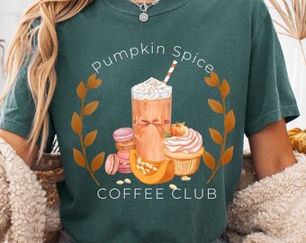 May include: Teal green t-shirt with a graphic that reads "Pumpkin Spice Coffee Club." The design features a pumpkin spice latte, macarons, and a cupcake. The shirt is worn by a person with blonde hair and blue jean shorts.