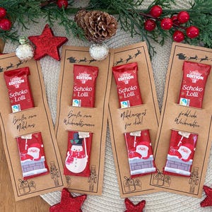 Puede incluir: Conjunto de regalo navide&ntilde;o con cuatro barras de chocolate en envoltorios rojos, cada una unida a una tarjeta marr&oacute;n con saludos festivos. La escena incluye decoraciones de estrellas rojas, vegetaci&oacute;n y una estera tejida. Las barras de chocolate tienen dise&ntilde;os festivos.