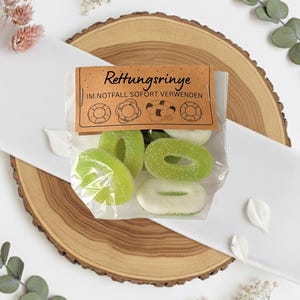 Puede incluir: Una bolsa transparente llena de caramelos de goma verdes y blancos con forma de salvavidas, con una etiqueta marrón que dice "Rettungsringe" y "IM NOTFALL SOFORT VERWENDEN". Los caramelos están sobre una rodaja de madera y tela blanca.