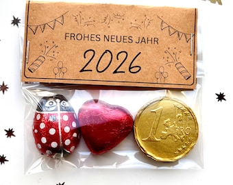 May include: A clear cellophane bag contains a ladybug-shaped chocolate, a heart-shaped chocolate, and a gold-colored chocolate coin. A brown paper tag reads "FROHES NEUES JAHR 2026". Scattered around are star-shaped confetti and "HAPPY NEW YEAR" decorations.