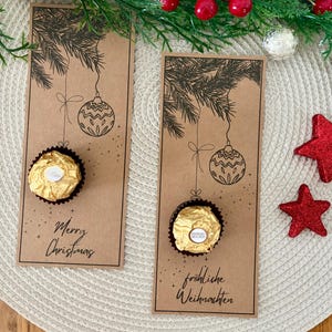 Può includere: Due biglietti regalo natalizi marroni con un disegno a tratto nero di un ornamento natalizio e rami di pino. Un biglietto dice "Merry Christmas" e l'altro dice "fröhliche Weihnachten". Ogni biglietto contiene un cioccolato dorato. Stelle rosse glitterate e verde sullo sfondo.