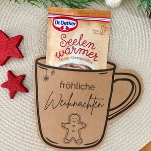 Könnte beinhalten: Ein weihnachtliches Bild mit einem braunen Pappbecherhalter mit den Worten "fröhliche Weihnachten" und einer Lebkuchenmann-Illustration. Ein Päckchen Dr. Oetker "Seelenwärmer" befindet sich darin. Rote Sternornamente und Grünzeug vervollständigen die Urlaubsszene.
