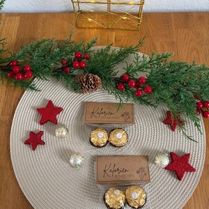 May include: A Christmas-themed display with two boxes of chocolates, red star decorations, and a garland with red berries and pine cones. The boxes are labelled "Kalorien Bomchen". The arrangement is on a beige placemat.