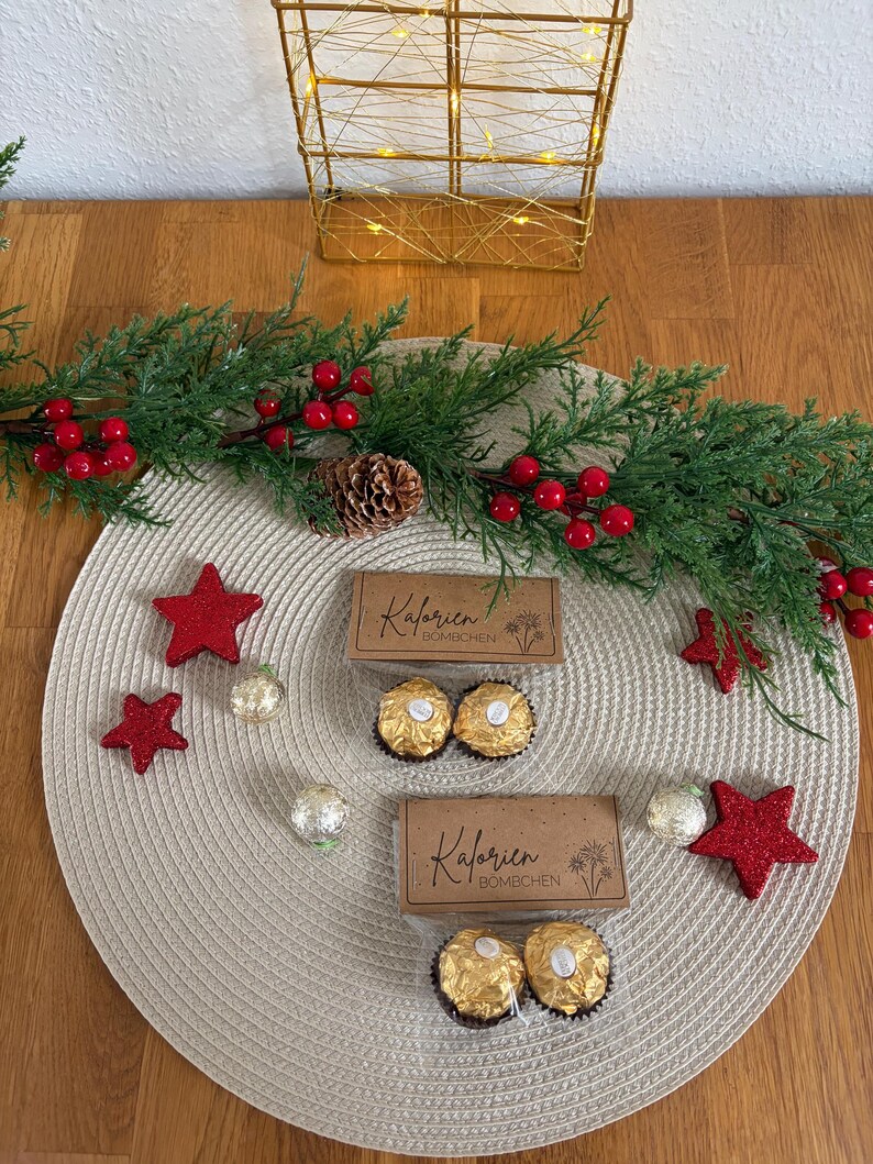 May include: A Christmas-themed display with two boxes of chocolates, red star decorations, and a garland with red berries and pine cones. The boxes are labelled "Kalorien Bomchen". The arrangement is on a beige placemat.