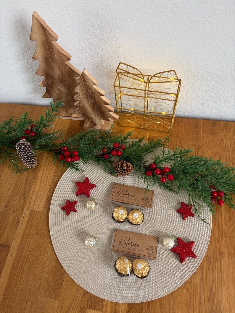 May include: A Christmas-themed display with wooden tree decorations, a gold wire gift box with fairy lights, and a garland with red berries and pine cones. Two boxes of chocolates, red star ornaments, and decorative spheres are also present.