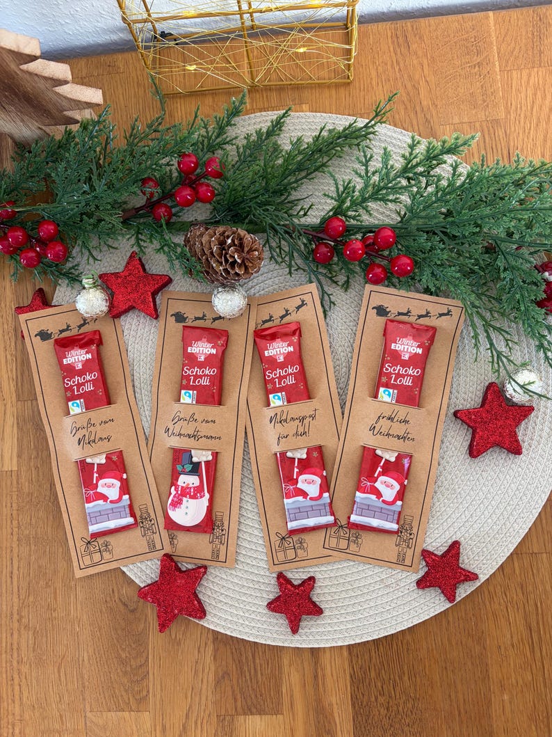 Puede incluir: Conjunto de regalo navide&ntilde;o con cuatro barras de chocolate en envoltorios rojos, cada una unida a una tarjeta marr&oacute;n con saludos festivos. La escena incluye decoraciones de estrellas rojas, vegetaci&oacute;n y una estera tejida. Las barras de chocolate tienen dise&ntilde;os festivos.