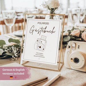 May include: A gold-framed sign with the word "POLAROID" and "Gästebuch" in elegant script. The sign features a line drawing of a camera and text in German. A white instant camera sits to the right, suggesting a guest book theme.