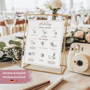 May include: A gold-framed sign with German instructions for a camera, next to a white instant camera. The sign details battery and film replacement. A pink banner at the bottom indicates German and English versions are included.