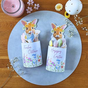 Könnte beinhalten: Zwei Osterdekorationen mit Hasenfiguren mit Blumenkränzen. Auf einer steht "Frohe Ostern" und auf der anderen "Happy Easter" mit Eiern und Blumen. Die Dekorationen befinden sich auf einer grauen kreisförmigen Oberfläche.