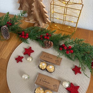 May include: A Christmas-themed display with two boxes of chocolates, red star decorations, and evergreen foliage. Wooden Christmas trees and a gold wire gift box with lights add to the festive scene, creating a warm and inviting atmosphere.