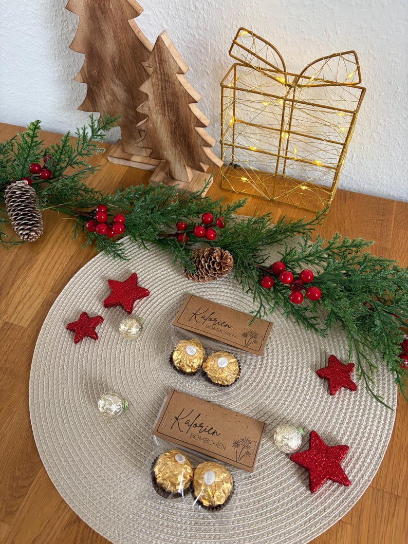 May include: A Christmas-themed display with two boxes of chocolates, red star decorations, and evergreen foliage. Wooden Christmas trees and a gold wire gift box with lights add to the festive scene, creating a warm and inviting atmosphere.
