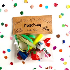 Puede incluir: Una bolsa de celofán transparente llena de obsequios para fiestas, que incluyen un marcador amarillo, una cinta verde y caramelos envueltos. Una etiqueta de papel marrón con la palabra "Fasching" está adjunta, rodeada de confeti colorido.