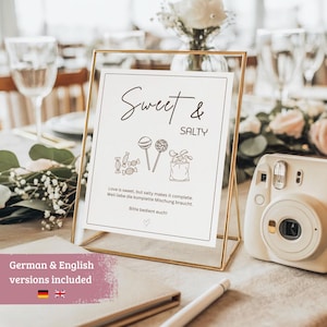 Könnte beinhalten: Ein goldgerahmtes Schild mit den Worten "Sweet & Salty" in eleganter Schrift, mit Süßigkeiten-Illustrationen. Das Schild enthält Text auf Englisch und Deutsch und steht auf einem Tisch mit einer Kamera und Blumengestecken.