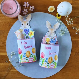 May include: Two Easter-themed paper crafts with bunny and spring flower designs. One says "Frohe Ostern" and the other says "Happy Easter". The crafts are on a gray circular surface, with a candle and decorative flowers in the background.