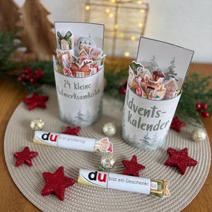 Könnte beinhalten: Zwei Adventskalender-Behälter aus Glas mit festlichen Dekorationen. Jeder Behälter hat ein gedrucktes Etikett und ist mit kleinen, weihnachtlichen Ausschnitten gefüllt. Rote Glitzersterne und Schokoriegel mit deutschem Text sind verstreut.