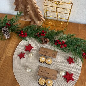 May include: A Christmas-themed display with wooden trees, a gold wire gift box with lights, and a garland with red berries and pinecones. Two boxes of chocolate candies and red star decorations are on a beige placemat.