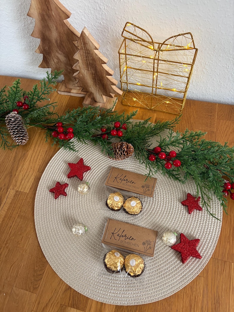May include: A Christmas-themed display with wooden trees, a gold wire gift box with lights, and a garland with red berries and pinecones. Two boxes of chocolate candies and red star decorations are on a beige placemat.