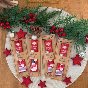Puede incluir: Chupetines de chocolate con tem&aacute;tica navide&ntilde;a en tarjetas decorativas. Cada tarjeta presenta un envoltorio rojo con el texto "Schoko Lolli" y una ilustraci&oacute;n festiva de Pap&aacute; Noel o un mu&ntilde;eco de nieve. Estrellas rojas brillantes y vegetaci&oacute;n rodean las tarjetas.