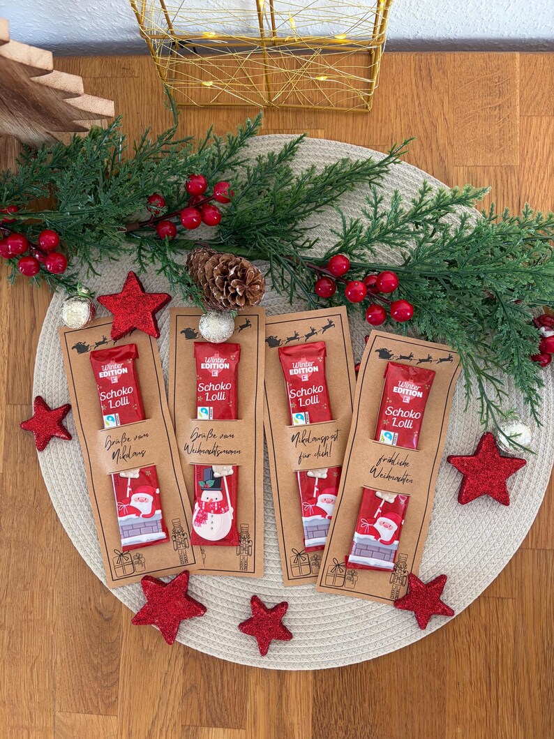Puede incluir: Chupetines de chocolate con tem&aacute;tica navide&ntilde;a en tarjetas decorativas. Cada tarjeta presenta un envoltorio rojo con el texto "Schoko Lolli" y una ilustraci&oacute;n festiva de Pap&aacute; Noel o un mu&ntilde;eco de nieve. Estrellas rojas brillantes y vegetaci&oacute;n rodean las tarjetas.