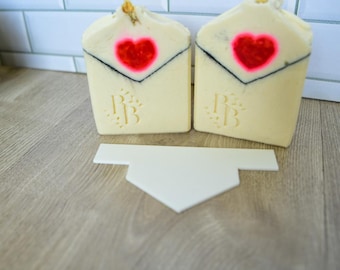 May include: Two white soap bars with a red heart design and the letters "BB" embossed on the front. The soap bars are sitting on a wooden surface.