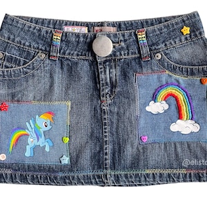 May include: A denim skirt with rainbow embroidery. The skirt has two pockets, one with a rainbow and clouds and the other with a rainbow and a cartoon pony. The skirt has a button closure and a zipper.