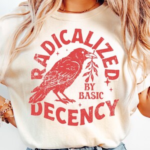 以下が含まれることがあります： クリーム色のTシャツに、赤色のかすれたグラフィック。グラフィックは小枝をくわえたカラスで、「RADICALIZED」の文字が上に、「BY BASIC DECENCY」の文字が下に配置されています。明るい色のブルージーンズと合わせています。