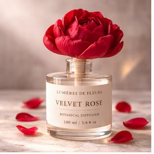 Puede incluir: Un difusor botánico de vidrio transparente con una gran rosa roja de terciopelo. La etiqueta dice "LUMIERES DE FLEURS VELVET ROSE BOTANICAL DIFFUSER 100 ml". Pétalos de rosa rojos esparcidos.