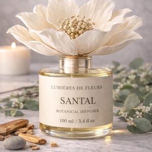 Puede incluir: Un difusor botánico de vidrio transparente con una gran flor de color crema en la parte superior. La etiqueta dice "SANTAL BOTANICAL DIFFUSER 100 ml / 3.4 fl oz." El difusor está rodeado de elementos decorativos, incluyendo trozos de madera y una vela encendida.