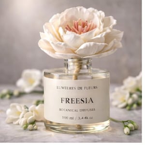 Puede incluir: Un difusor de vidrio transparente con una gran flor blanca en la parte superior. La botella tiene una etiqueta que dice "LUMIERES DE FLEURS" y "FREESIA BOTANICAL DIFFUSES 100 ml". La flor tiene un centro rosa. Flores blancas adicionales en el fondo.