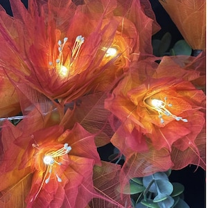 May include: Close-up of illuminated orange and red decorative flowers. The flowers are made of delicate, veined material, with small white and blue accents in the centre. The warm glow of the lights creates a vibrant and inviting display.