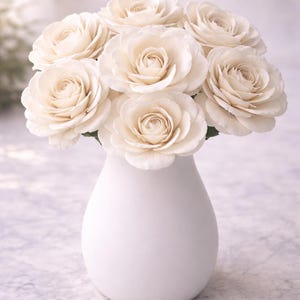 May include: A bouquet of ivory-colored roses in a white, teardrop-shaped vase. The roses have multiple layers of petals, and the vase has a smooth, matte finish. The arrangement is set against a soft, blurred background.