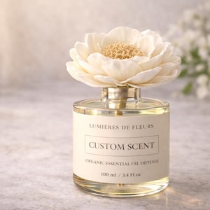 May include: A glass essential oil diffuser with a cream-coloured flower. The label reads "LUMIÈRES DE FLEURS CUSTOM SCENT ORGANIC ESSENTIAL OIL DIFFUSER 100 ml / 3.4 Fl oz."