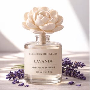 May include: A clear glass botanical diffuser with a cream-colored flower on top. The label reads "Lumières de Fleurs Lavande Botanical Diffuser 100 ml / 3.4 fl oz." Sprigs of lavender are arranged around the bottle.