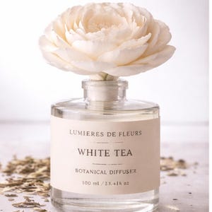 May include: A clear glass botanical diffuser with a large, cream-colored flower on top. The label reads "LUMIERES DE FLEURS WHITE TEA BOTANICAL DIFFUSER" and includes the volume: 100 ml / 3.4 fl oz. The diffuser is designed for home fragrance.