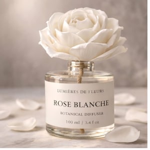 May include: A clear glass botanical diffuser with a large white rose on top. The label reads "LUMIÈRES DE FLEURS ROSE BLANCHE BOTANICAL DIFFUSER 100 ml / 3.4 fl oz". Scattered white rose petals surround the diffuser.