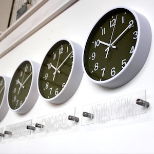 Puede incluir: Cuatro relojes de pared redondos con marcos blancos y esferas verde oliva están montados en una pared blanca. Los relojes muestran diferentes horas. Debajo de los relojes hay letreros de acrílico transparente con nombres de ciudades.