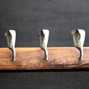 Puede incluir: Un expositor de palos de golf de madera oscura, con tres palos de golf plateados fijados a una base rectangular. Se aprecia la veta de la madera.