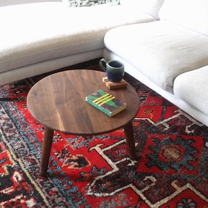 May include: A round, dark brown wooden coffee table with three tapered legs. On the table surface sits a green book with yellow stripes and a dark blue ceramic mug resting on a small, square wooden coaster.
