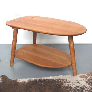 May include: A light brown wooden side table with an oval-shaped top and a matching oval-shaped lower shelf. The table features four tapered legs that extend outward from the center, showcasing a natural wood grain finish throughout the structure.