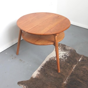 May include: A round wooden side table featuring a smooth, circular top and a smaller circular lower shelf. The table is supported by three tapered legs, each with subtle horizontal grooved detailing near the base, finished in a warm brown tone.
