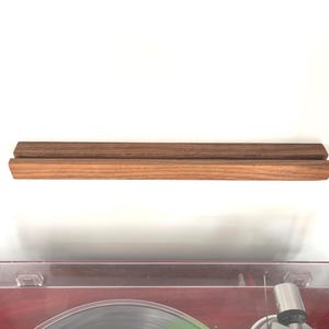Puede incluir: Un soporte de discos de madera, con dos piezas rectangulares paralelas. La pieza superior es de color marrón oscuro, mientras que la inferior es de color marrón rojizo claro. Diseñado para exhibir discos de vinilo. Mide unos 30 cm de largo.