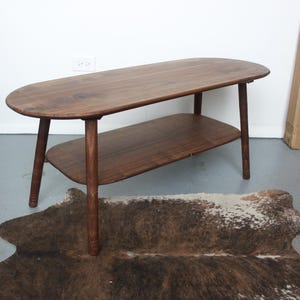 May include: A dark brown wooden coffee table with an oval-shaped top and a matching lower shelf. The table features four tapered legs and a smooth, polished finish, showcasing the natural wood grain patterns throughout the entire structure.