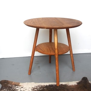 May include: A round wooden side table featuring a smooth, circular top and a smaller circular lower shelf. The table is supported by three tapered legs, each with subtle horizontal ring detailing near the base, showcasing a natural wood grain.