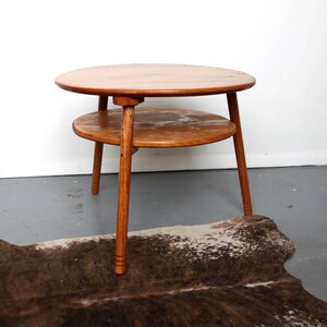 May include: A round wooden side table with a smooth, light brown finish. The table features a circular top and a smaller circular lower shelf supported by three tapered legs. The wood grain is visible across the surfaces of the table.