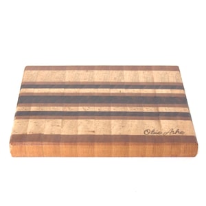 May include: A rectangular wooden cutting board featuring a striped pattern of light, medium, and dark brown wood grains. The surface displays a smooth finish with the text Obie Ashe engraved in the bottom right corner, showcasing a multi-toned design.