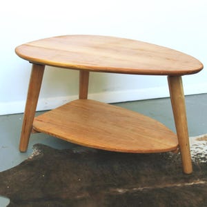 May include: A mid-century style wooden side table with an unusual, organic form. The table has a triangular top and a lower shelf, supported by three angled legs. The wood has a warm, natural finish. The table is approximately 76 cm long.