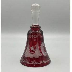 Puede incluir: Una campana de cristal rojo rubí con un asa de cristal transparente y complejos diseños grabados en blanco. El cuerpo de la campana muestra un patrón repetitivo de arcos y motivos florales en los bordes, con una figura de ave grabada.